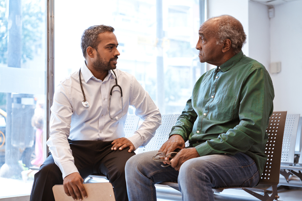 Doctor talking to senior patient sitting in waiting room. Serious man is staring at healthcare worker at hospital. They are against window.