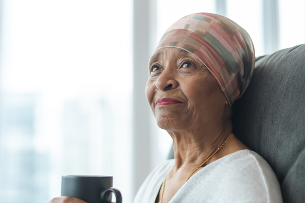 A black senior woman with cancer is wearing a scarf on her head. She is sitting at home in her living room and is holding a cup of tea. She is smiling thoughtfully while looking out a window. She is full of gratitude and hope for recovery.