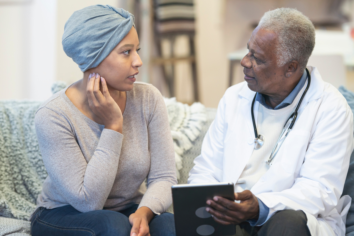 A mature adult physician is meeting with a patient in the patient's home. The patient is a black woman with cancer. The patient is wearing a headscarf to hide hair loss from chemotherapy treatment. The doctor and patient are seated next to each other on a couch. The doctor is holding a wireless tablet computer. The concerned patient is asking the doctor a question.