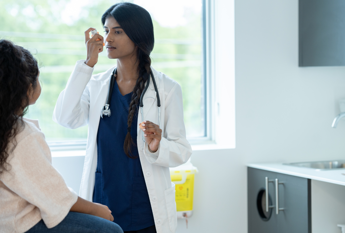 A female doctor holds out an inhaler as she instructs her young patient on how to properly use it for her Asthma. The patient is dressed casually and sitting up on the table as she pays close attention to the instructions.